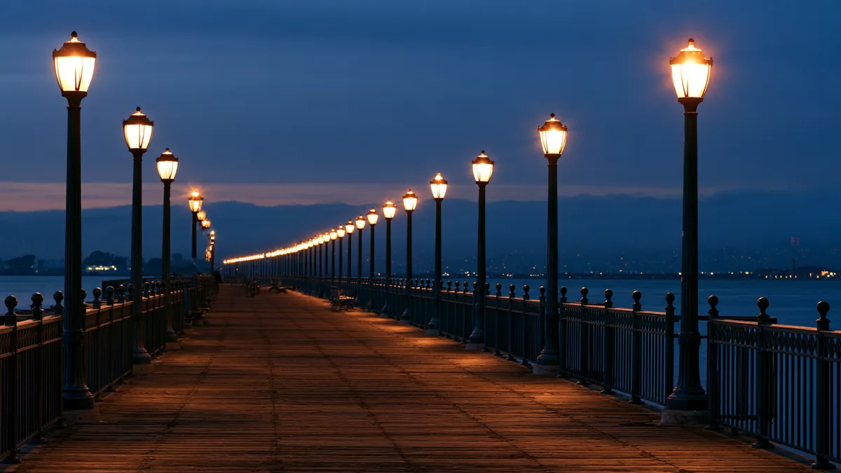 Symbolbild: Uferpromenade zur blauen Stunde mit einer langen Reihe warm leuchtender Laternen, die sich bis zum Horizont fortsetzt